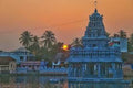 A South Indian Hindu temple with a blue-gray Dravidian-style tower stands in the middle of a calm temple tank at sunset. The orange sky glows behind the temple, with the sun setting near the horizon. Coconut palm trees and smaller temple buildings surround the water, and the temple's reflection is visible on the surface, creating a peaceful and scenic atmosphere.
