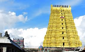 A tall, multi-tiered yellow Hindu temple tower (gopuram) under a bright blue sky with white clouds, with smaller structures visible to the left.